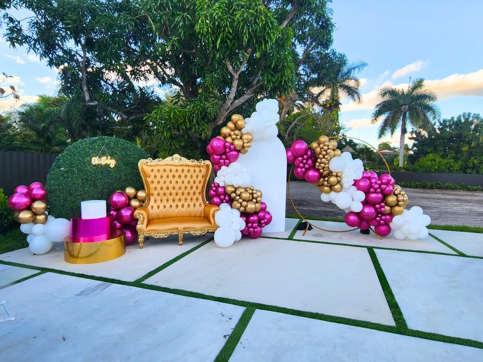 Outdoor balloon setup with a gold throne, magenta and gold garlands and a greenery wall