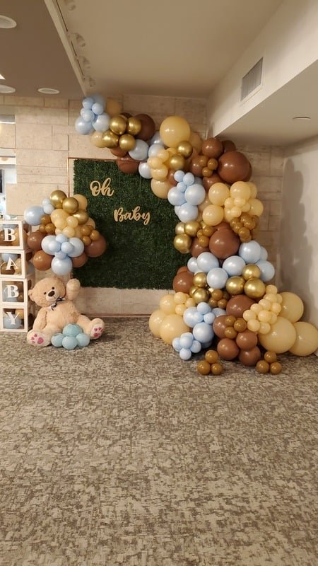 Brown, tan and blue baby-shower balloon garland on a greenery wall with a teddy bear